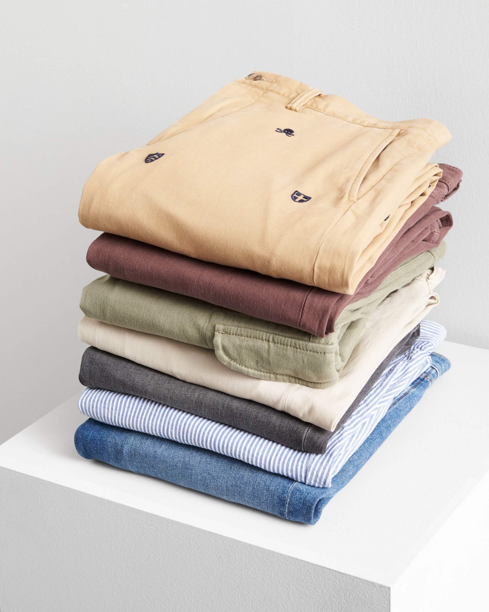 Stitch Fix men's stack of slim pants in denim, olive, cream, burgundy and printed yellow pants on a white cube.