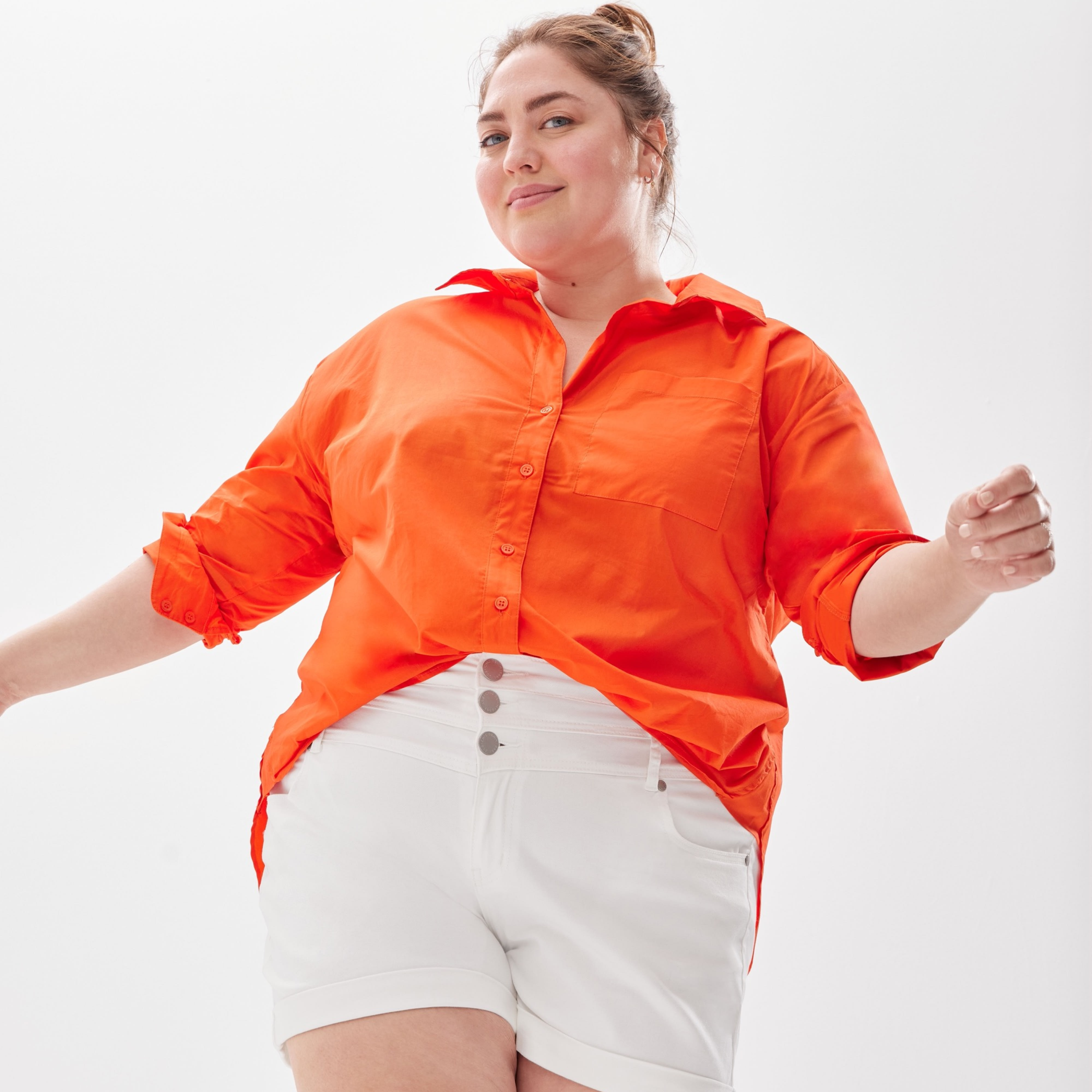 Plus-size model wearing women’s Stitch Fix clothing, including orange button-down shirt and white shorts.